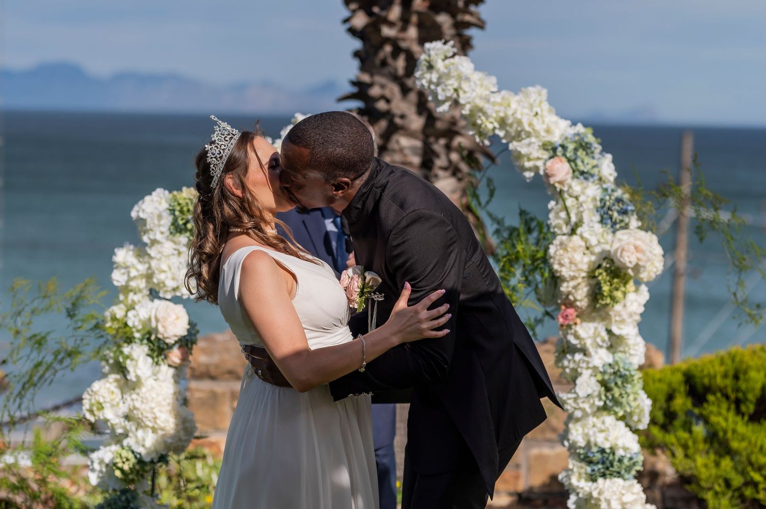 Couple sharing first kiss at outdoor beach wedding ceremony under floral arch with ocean backdrop.