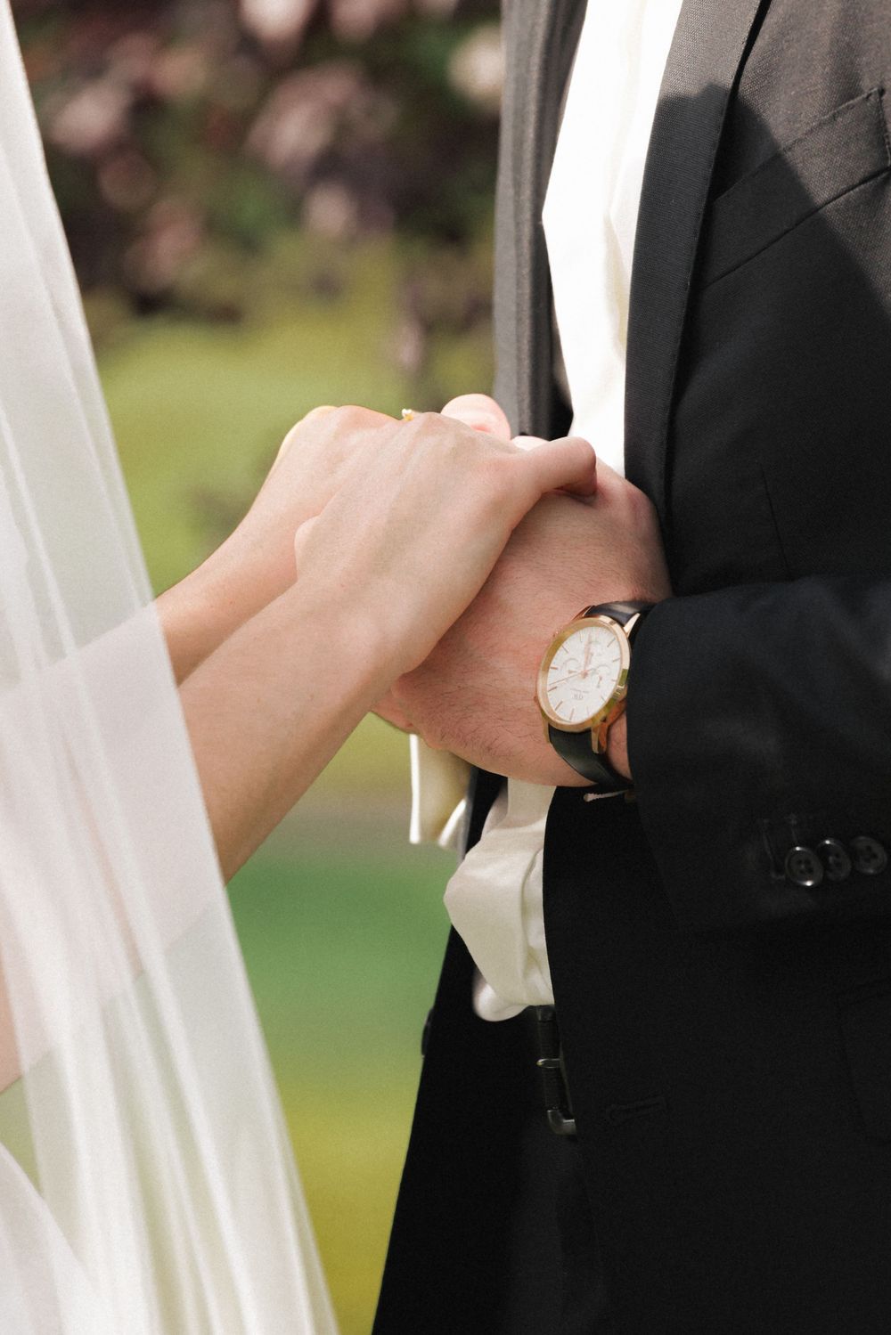 Bride and groom holding hands at wedding ceremony, groom wearing gold watch with black suit.