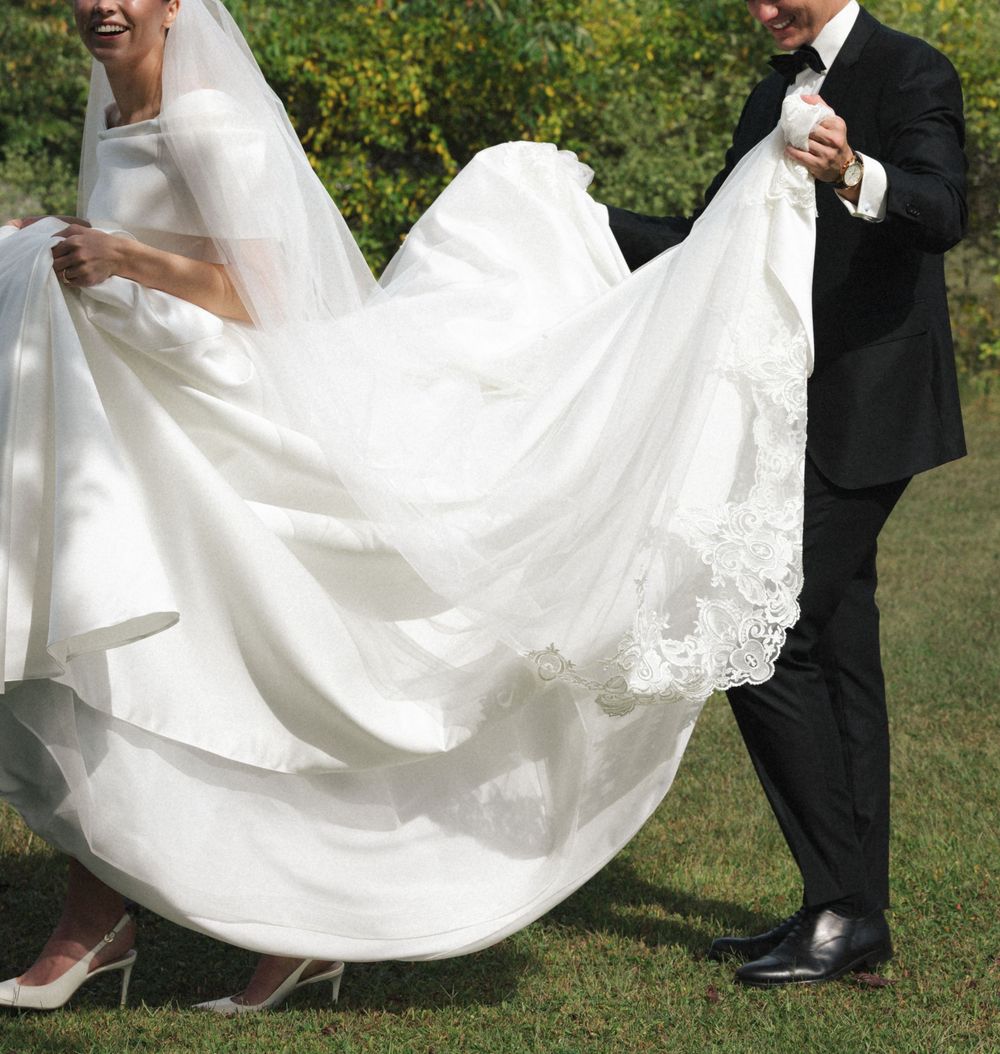 Bride in white lace wedding gown with veil, groom in black tuxedo holding dress train outdoors.