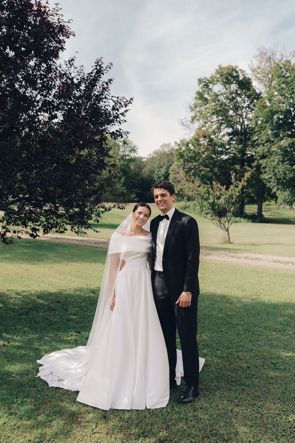 Smiling bride and groom pose together in a park, bride in white gown with veil, groom in black tuxedo.
