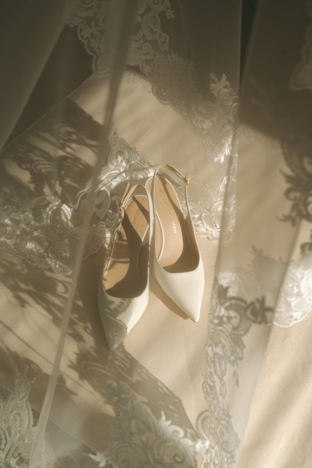 White pointed-toe bridal heels displayed against delicate lace wedding veil in soft sunlight.
