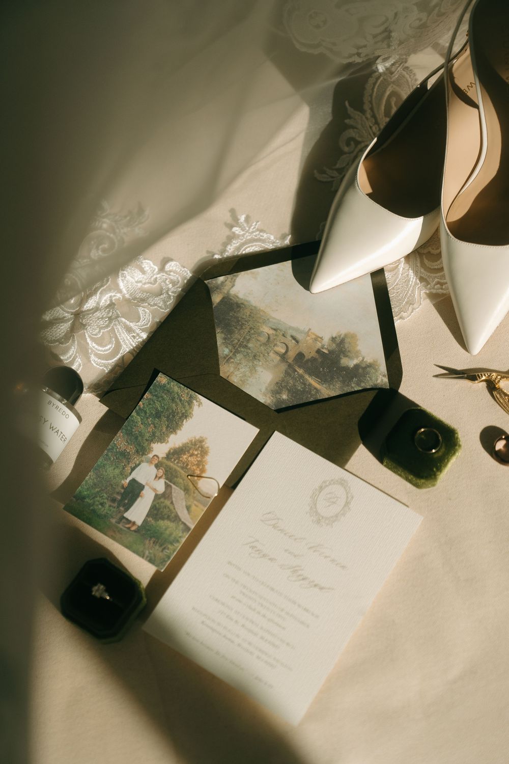 Flat lay of wedding invitation suite, white heels, rings, and accessories on lace.