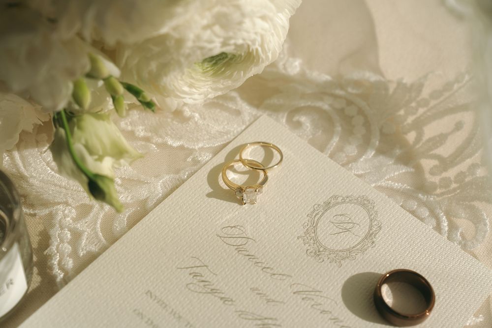 Elegant wedding flat lay with gold rings, white ranunculus flowers, and an ivory embossed invitation on lace fabric.