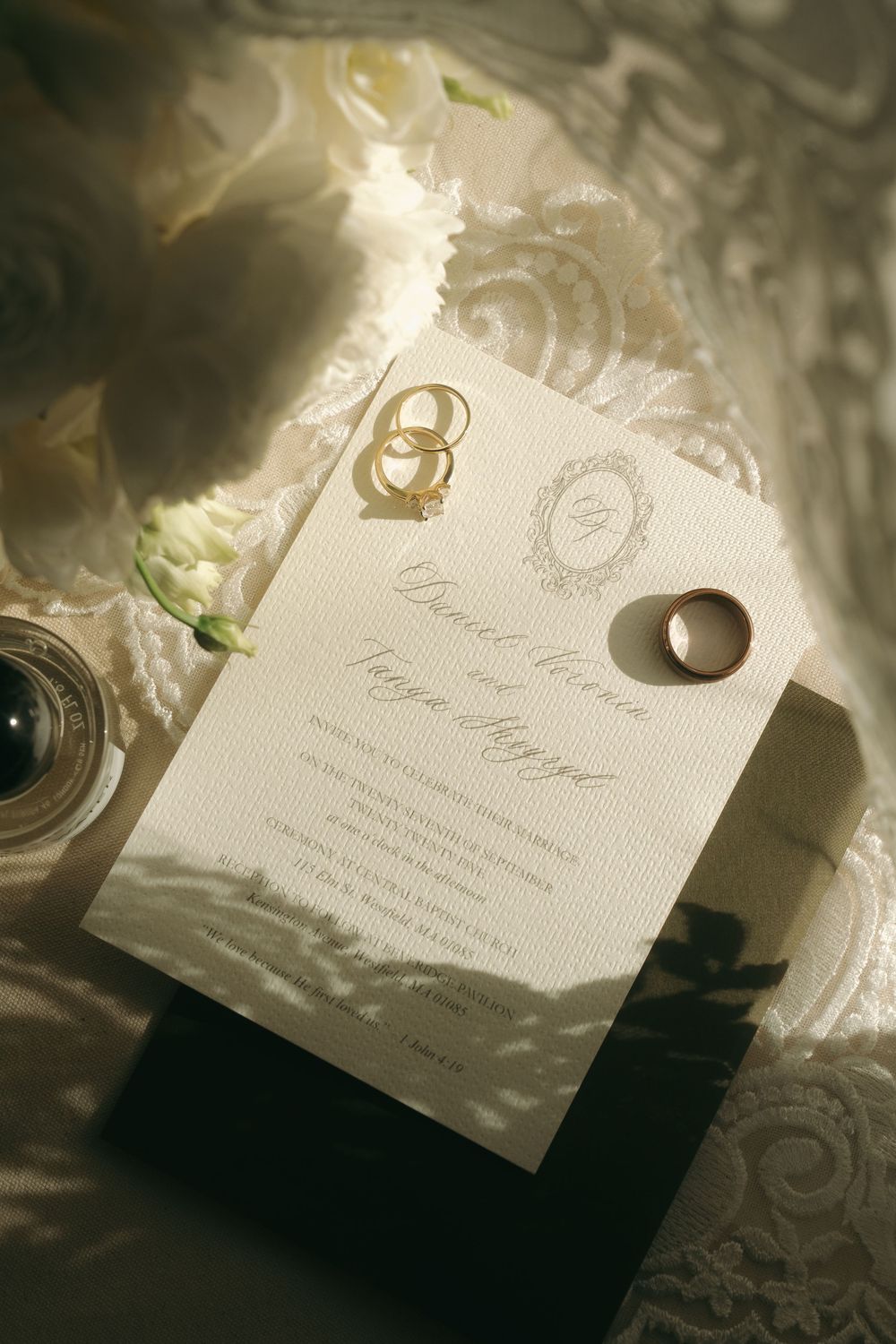 Elegant wedding invitation with gold rings and white floral arrangement on lace fabric in soft natural light.