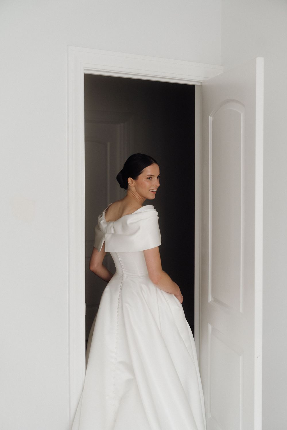 Smiling bride in off-shoulder white ball gown standing in white doorway looking back.