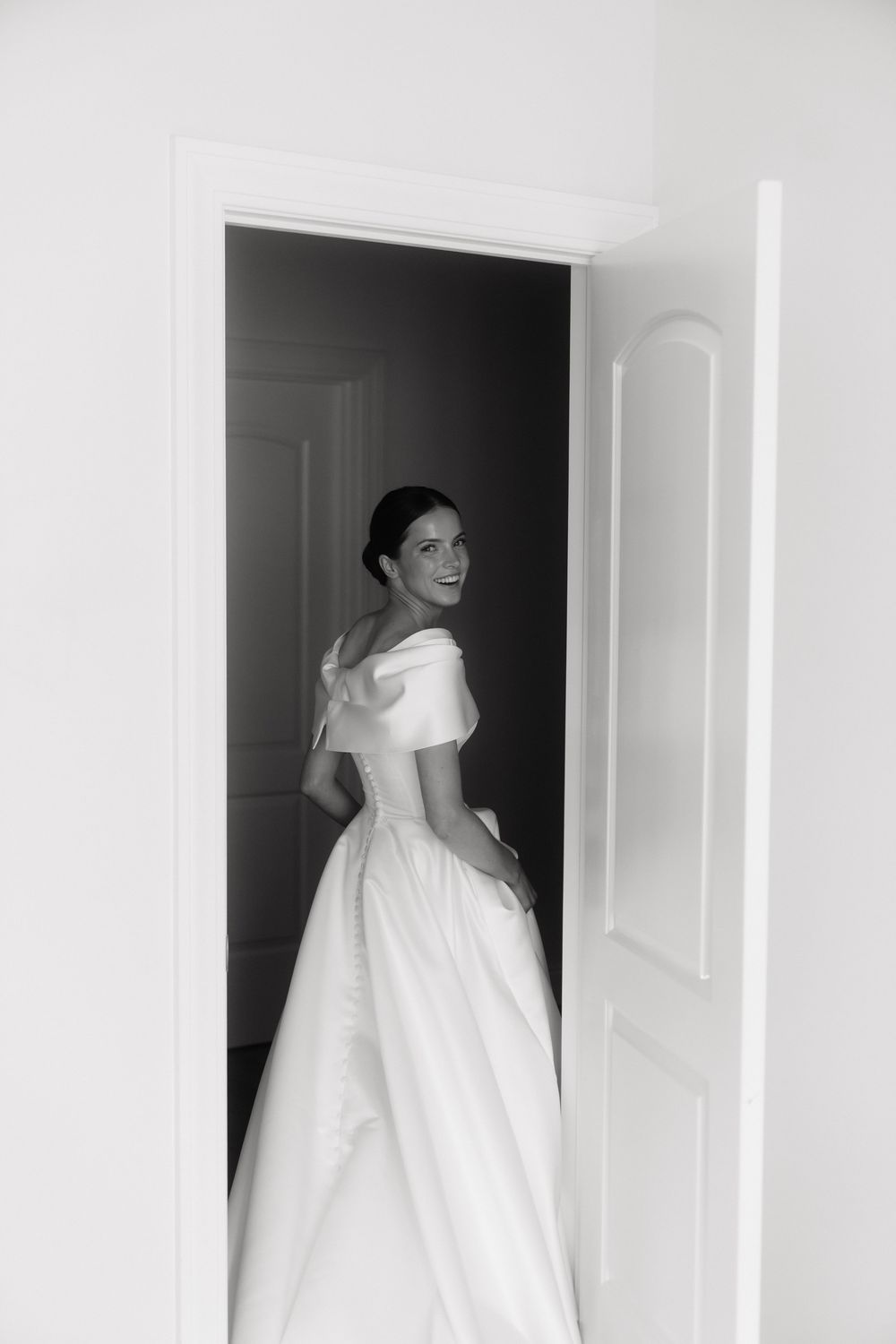Elegant bride in off-shoulder white ball gown smiling over shoulder in doorway, black and white wedding photo.