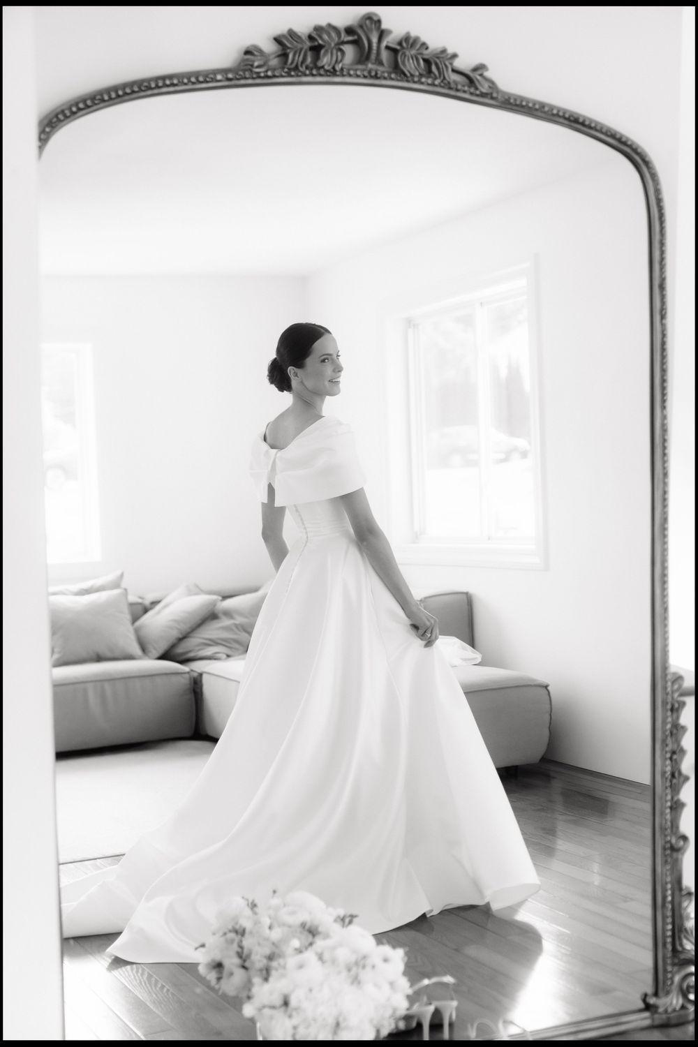 Bride in elegant off-shoulder white gown reflected in ornate gold mirror in bright white room.