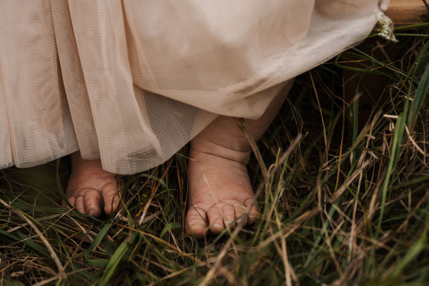 Bare feet peek out from beneath a flowing beige dress while standing in tall grass.