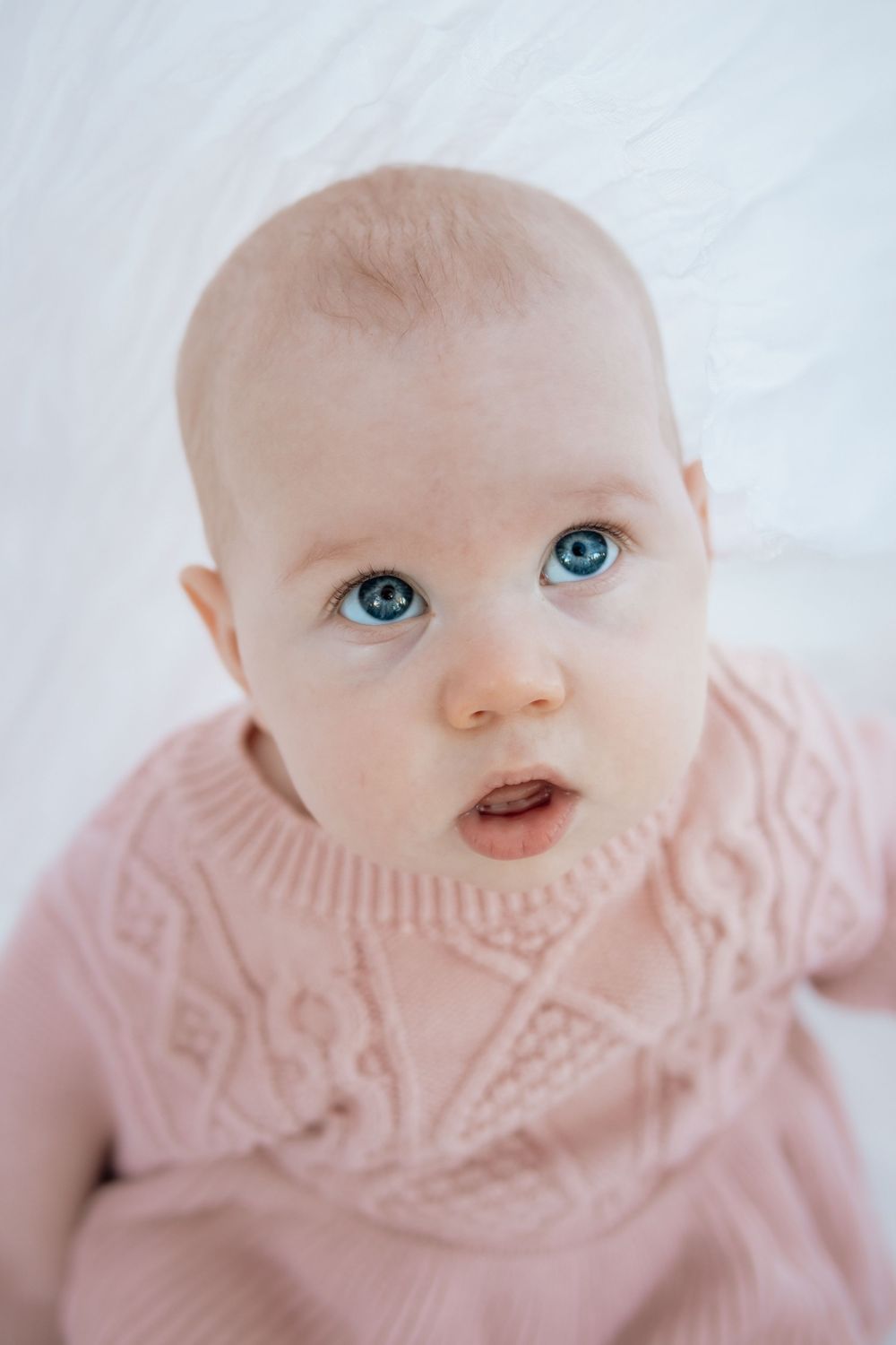 A baby wearing a pink knit sweater gazes upward with bright blue eyes against a light background.