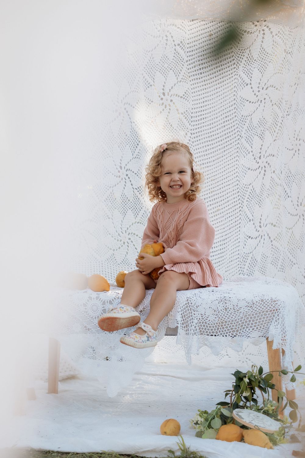Child in pink dress sits on white bench with citrus fruits and lace curtain backdrop in dreamy portrait setting.