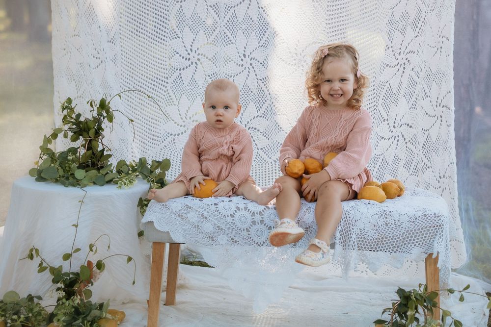 A group of children in white lace dresses sit together against a lace backdrop for a portrait session.