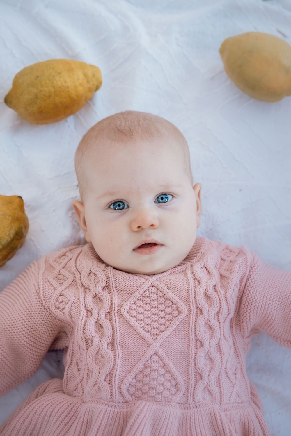 A baby wearing a pink knitted sweater with intricate cable patterns lies on a white surface.