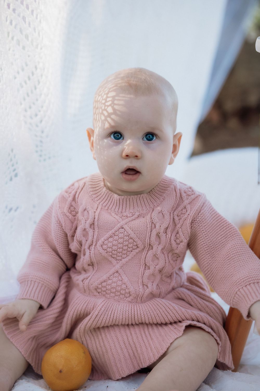 A baby wearing a pink knit sweater dress sits with an orange on a white blanket.