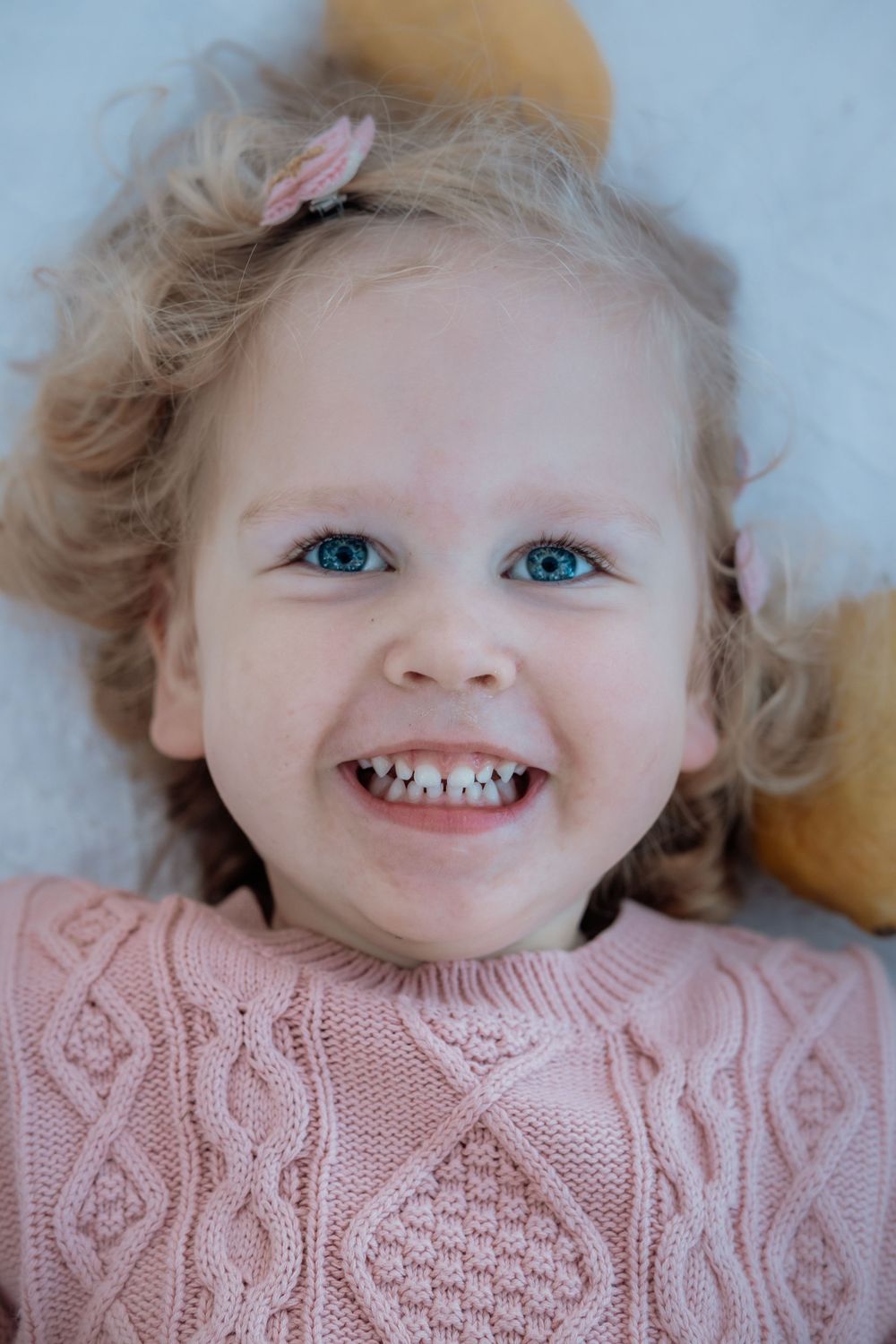 A young child wearing a pink knit sweater smiles brightly showing teeth with light blonde curly hair and blue eyes.