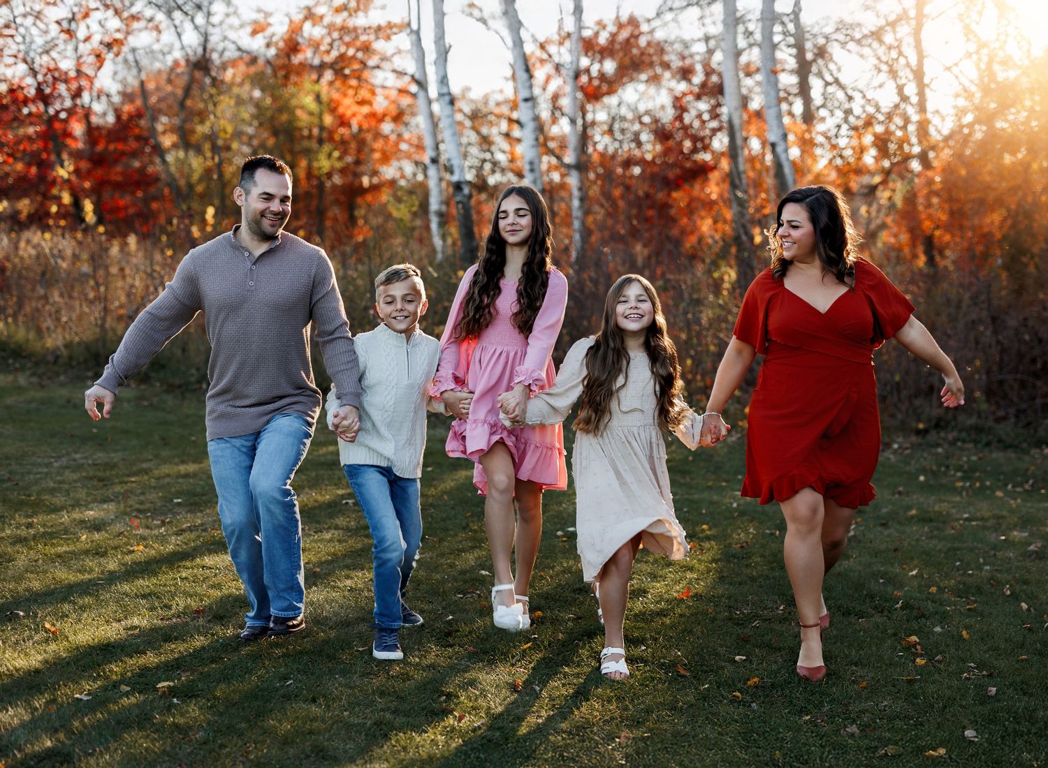 A family holds hands while walking together through an autumn forest at sunset with colorful fall foliage in background.
