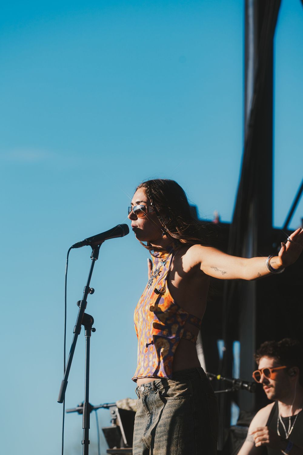 Performers on stage against bright blue sky with microphone stands silhouetted during outdoor concert performance.