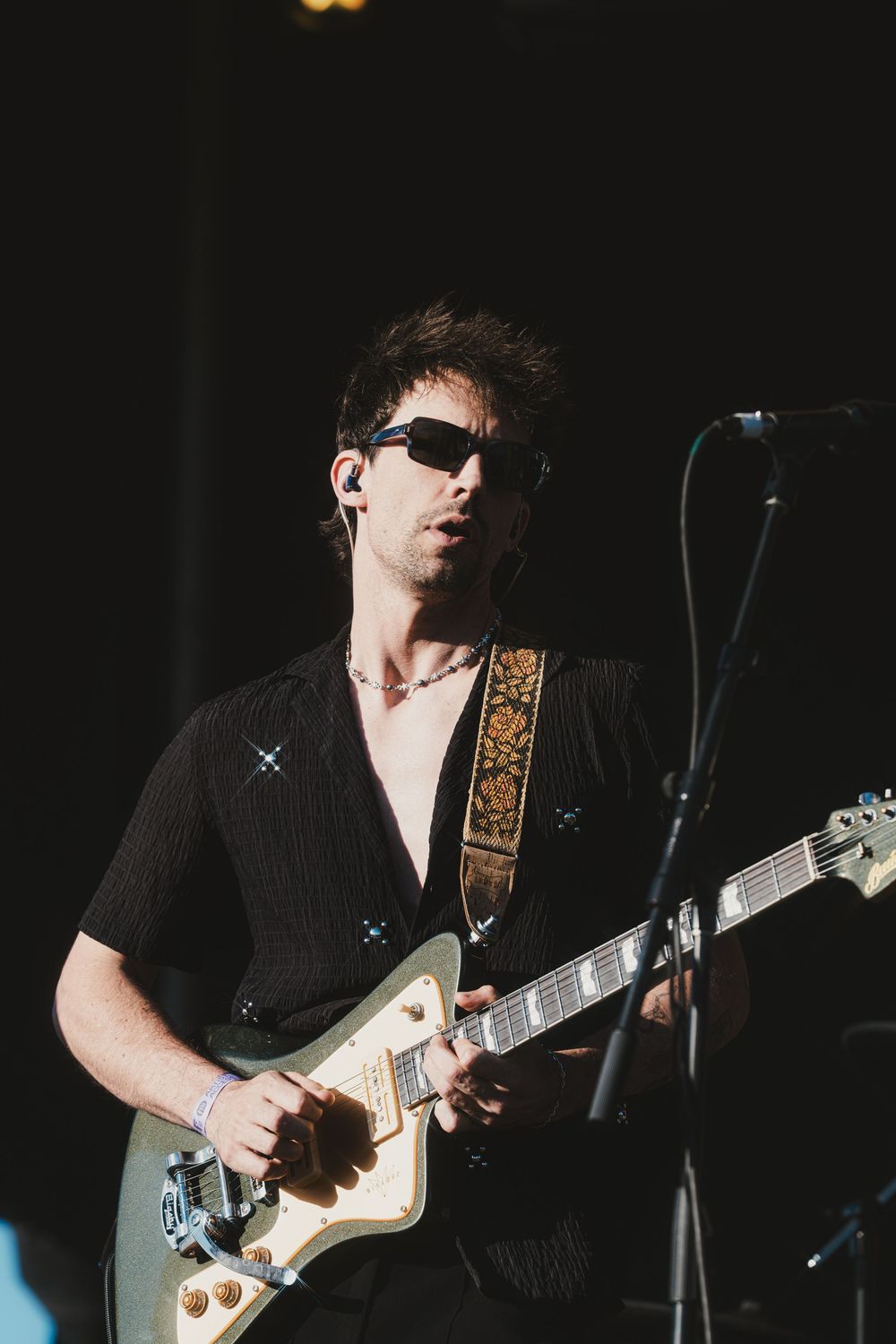 A guitarist performs on stage in dark lighting wearing black clothing and playing an electric guitar with a gold pickguard.