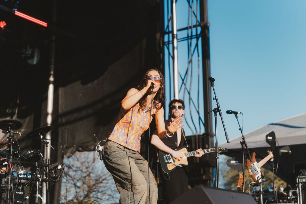 Musicians perform on stage with guitars and instruments during an outdoor concert at sunset.