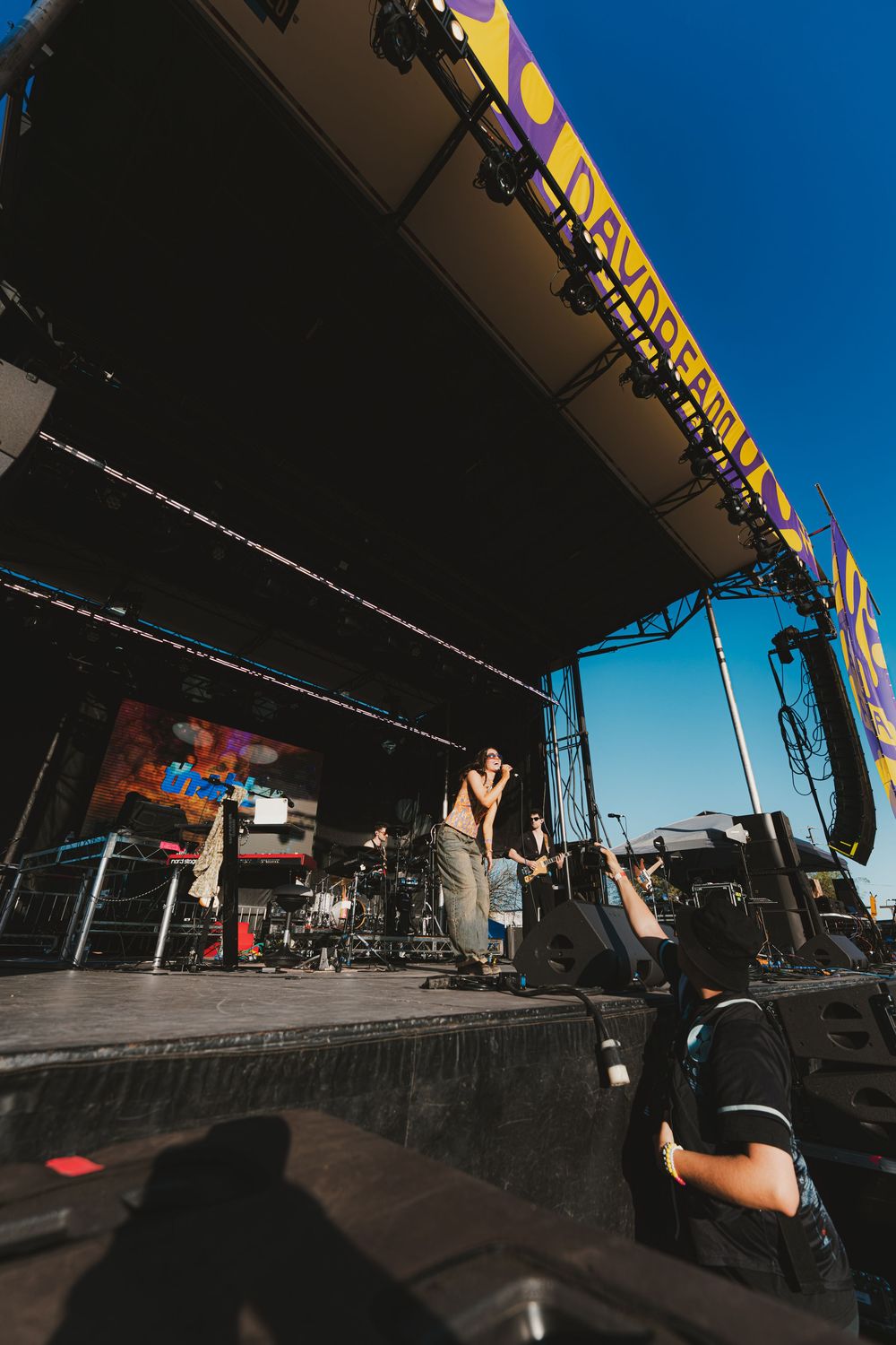 Wide angle view of an outdoor concert stage with equipment and performers under a bright blue sky.
