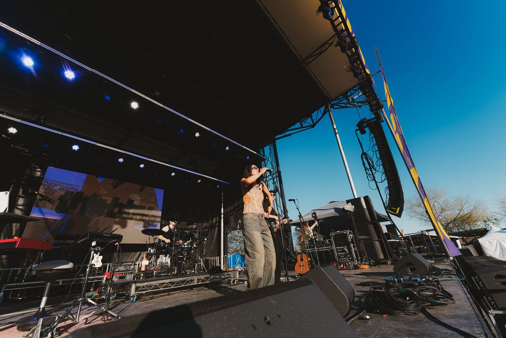 Wide angle view of an outdoor stage setup with lighting equipment and sound gear under a blue sky.
