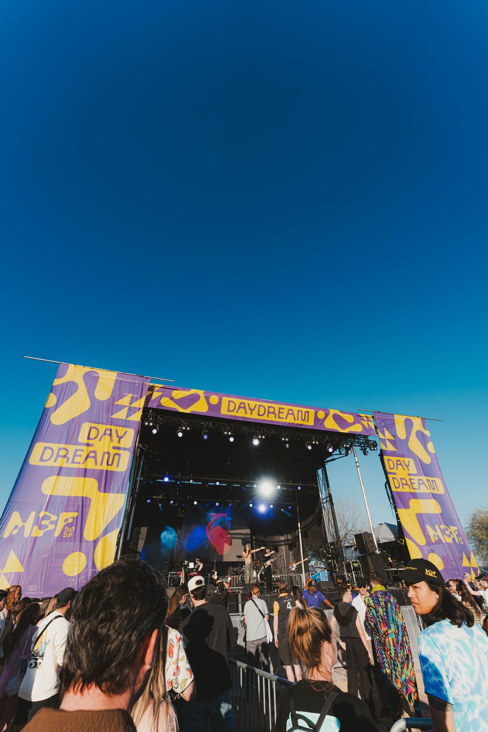 Concert crowd gathers at outdoor music festival stage with colorful purple banners and clear blue sky overhead.