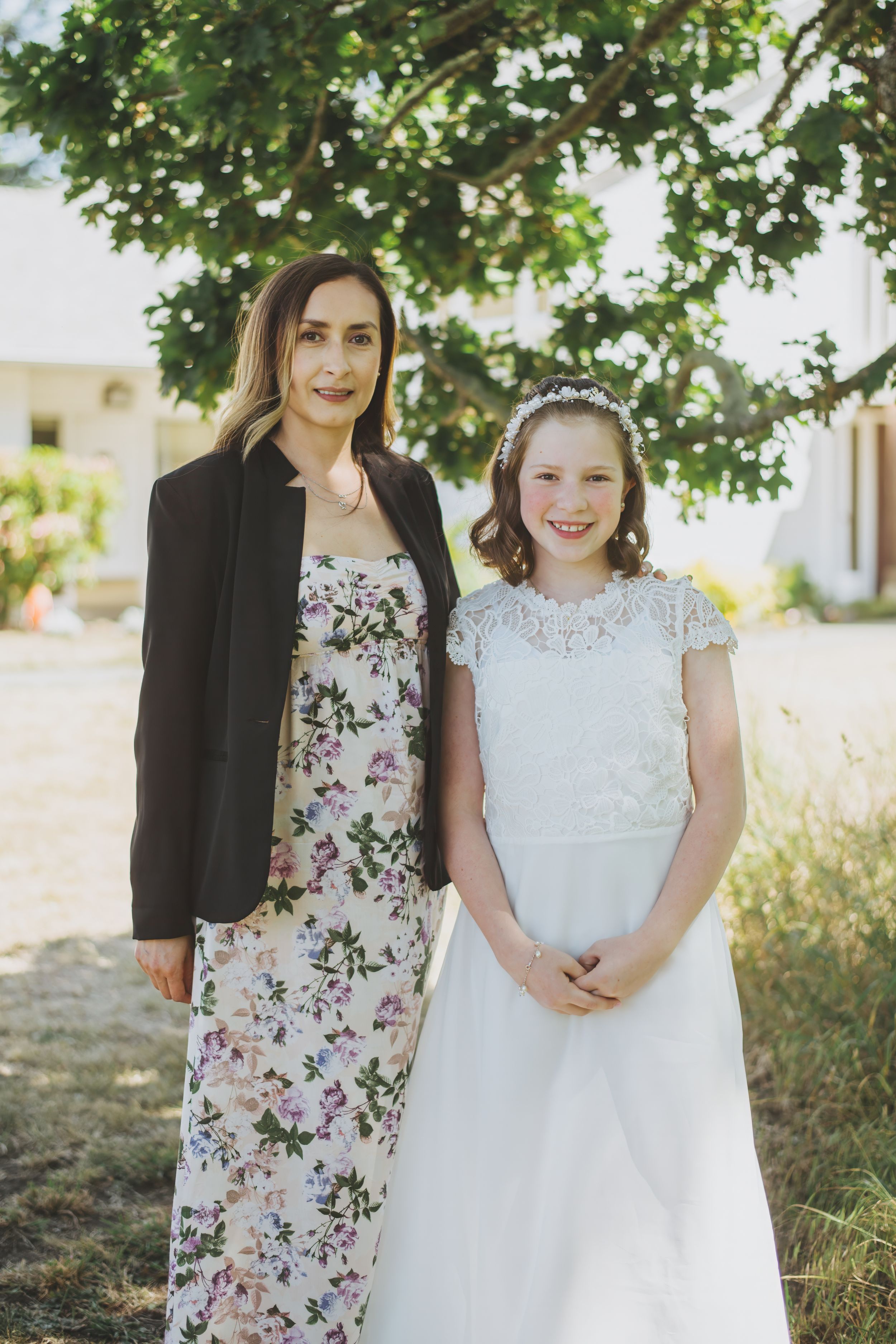 Two people pose outdoors in formal attire, one in a floral dress with black blazer and one in white lace dress.