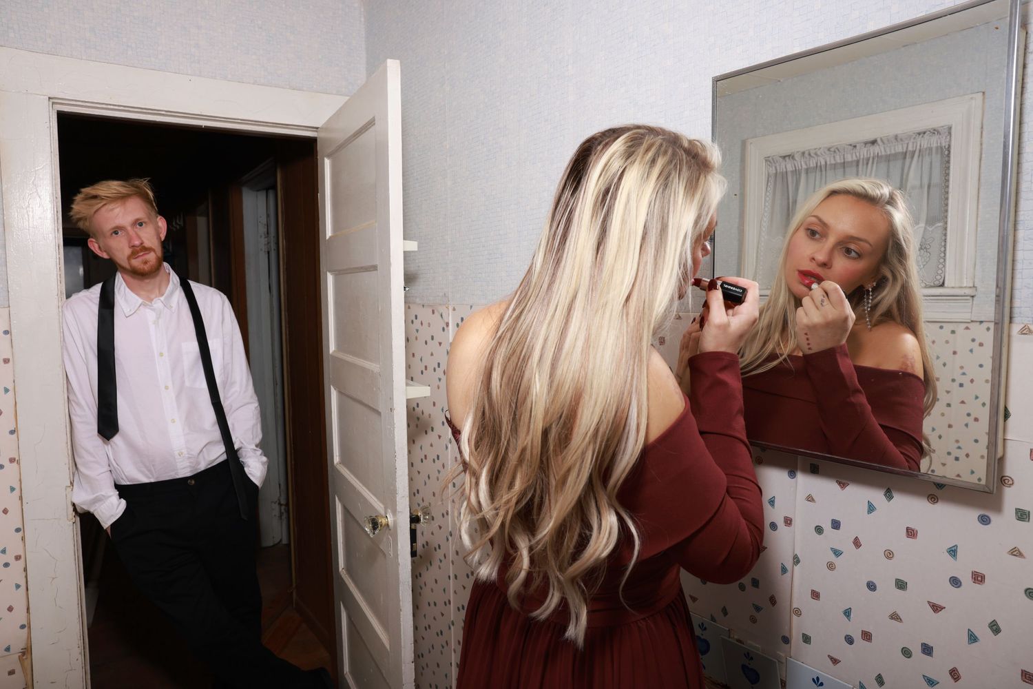 A person with blonde hair in a maroon shirt looks at their reflection in multiple wall mirrors.