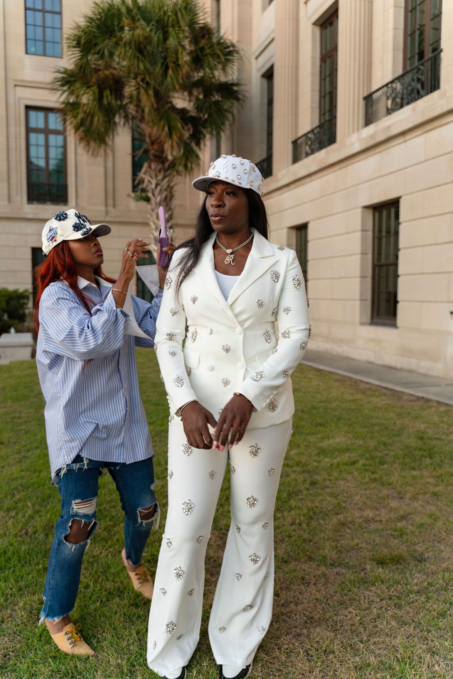 Two people pose playfully in casual outfits with white pants and blue tops against a building exterior with palm trees.