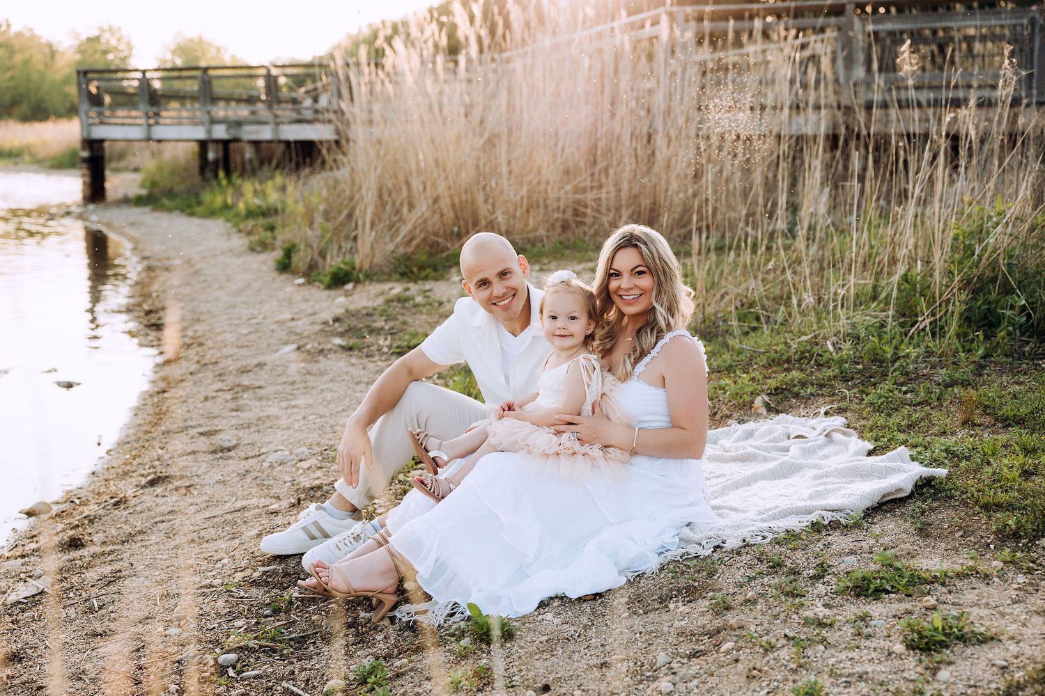 Beautiful warm evening session by the lake, with a warm sunset sun filling the image as the family of three sits on a blanket by the lake