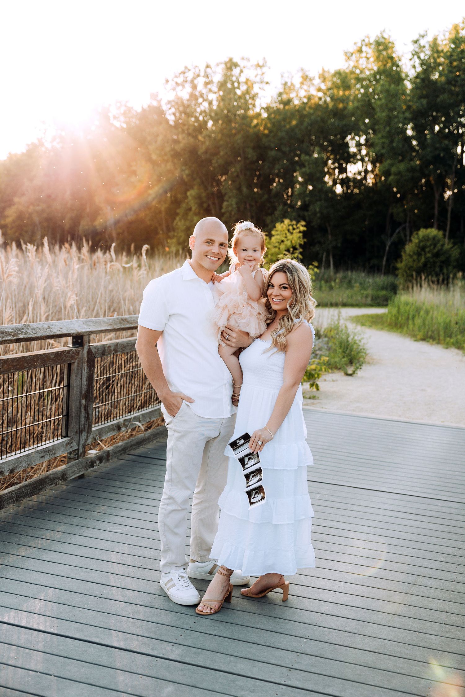 family of three, wearing white neutral outfits and mom holds an ultrasound picture for pregnancy announcement