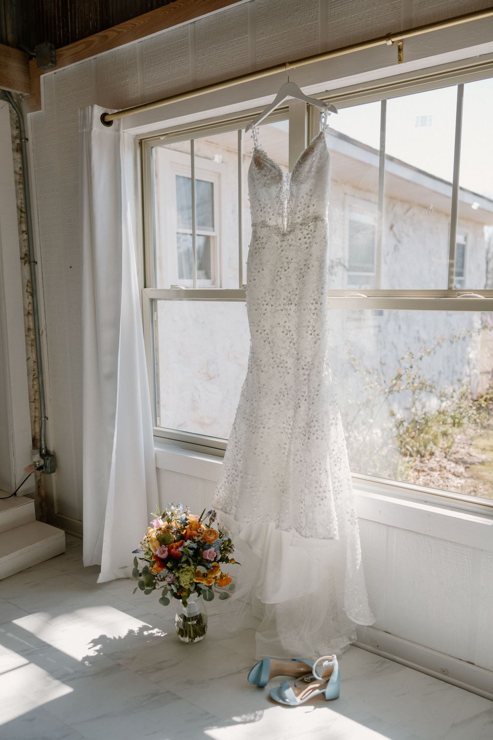 White wedding dress hanging in sunlit room with colorful floral arrangement below.