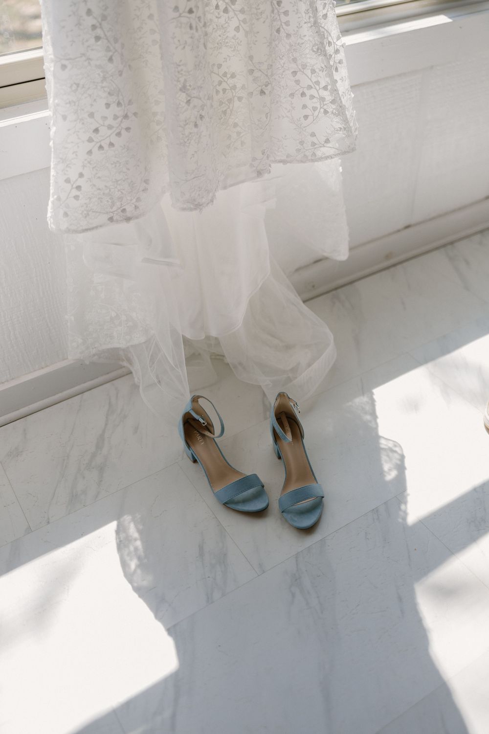 Light blue wedding shoes peek out from under a flowing white wedding dress near sunlit windows.