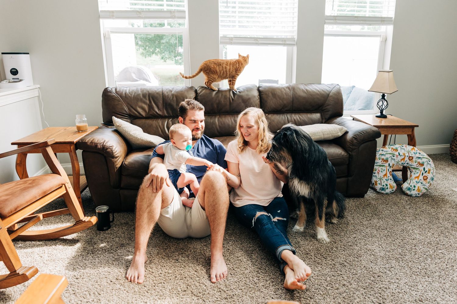 A couple relaxes on a leather couch in their living room with a ginger cat and black dog as pets and their baby.
