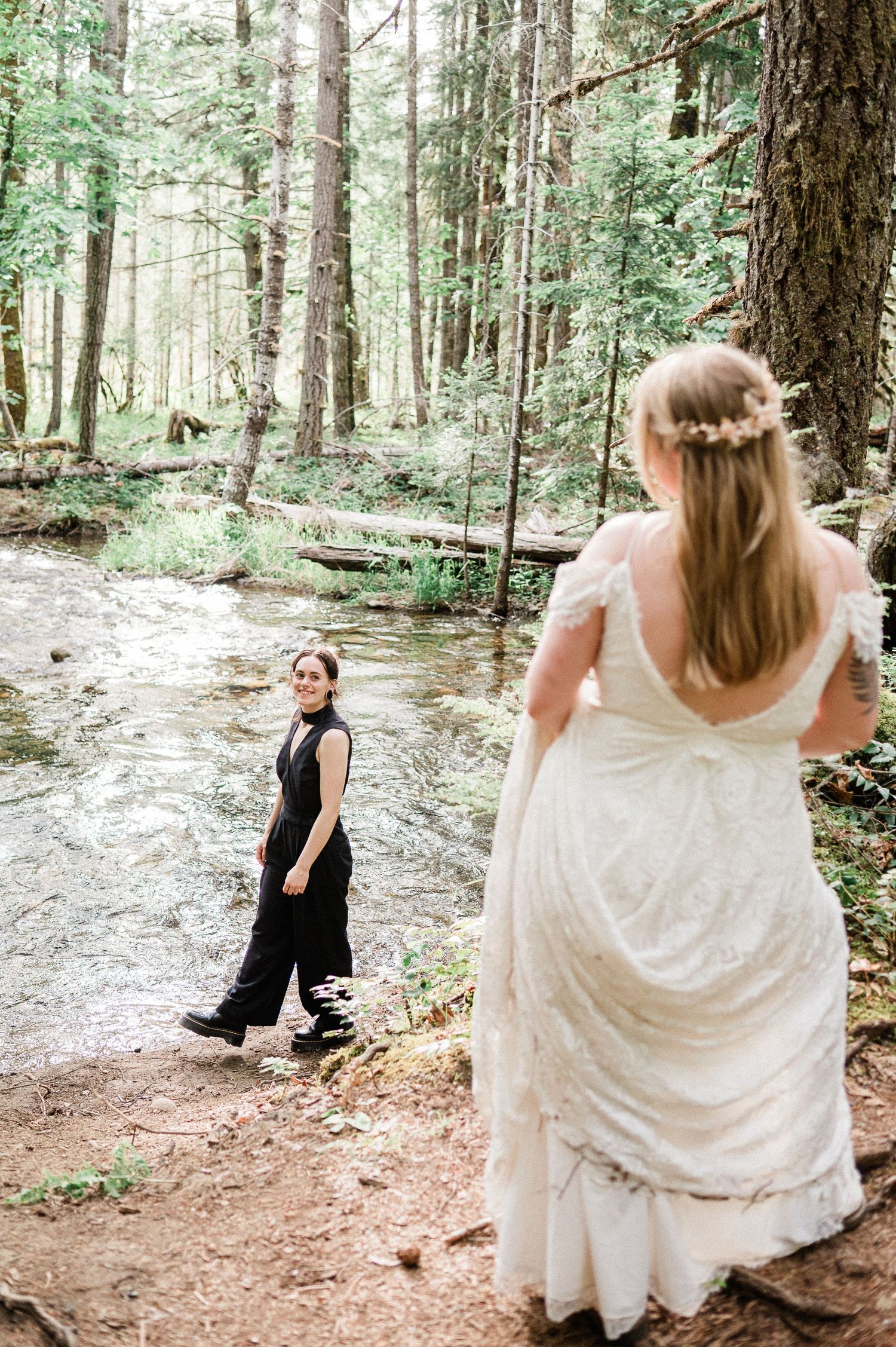 A couple in wedding attire playfully poses in a sunlit forest setting next to a flowing river.