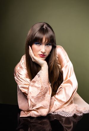 Girl in a pink robe in front of an olive backdrop, leaning over a reflective table, looking straight into camera
