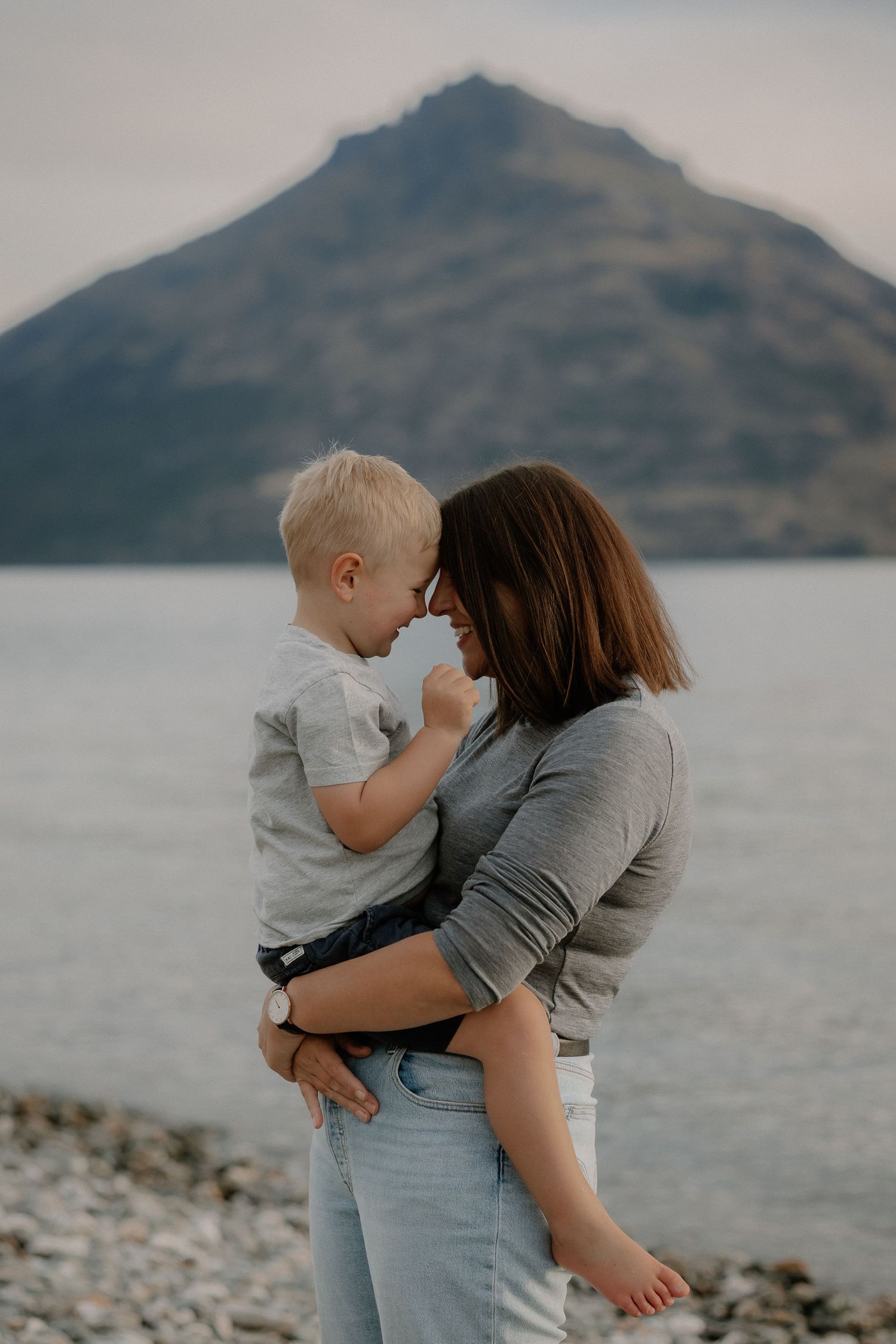 A mother holds her young blonde toddler close, touching noses by Lake Wakatipu, Homestead Bay in Queenstown during a family photoshoot.