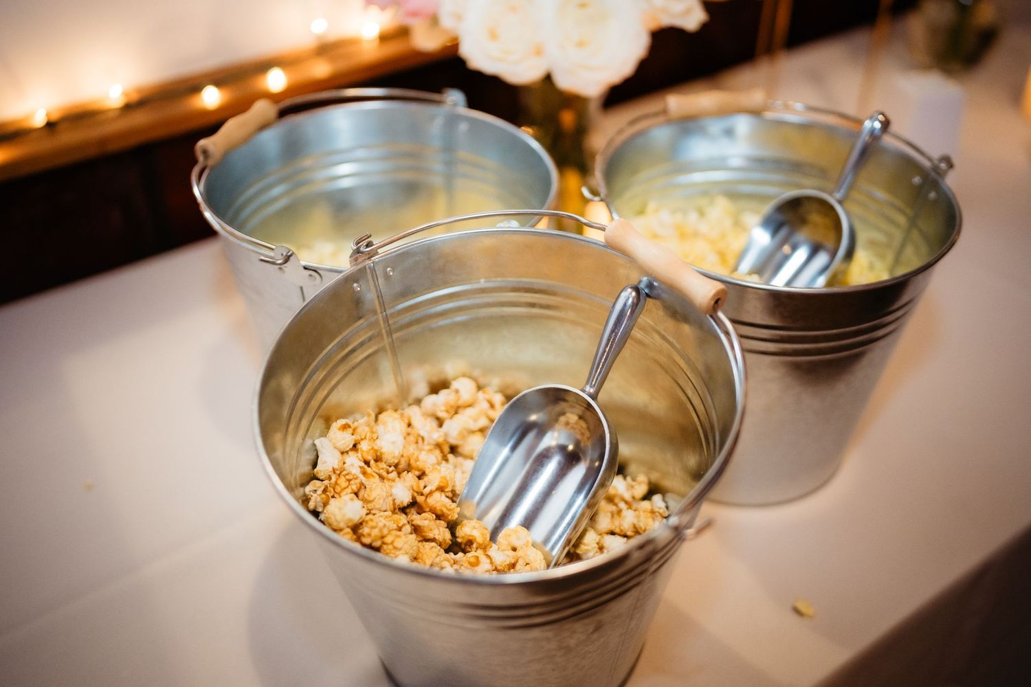 Three galvanized metal buckets filled with popcorn and scoops displayed on a white table at a catered event.