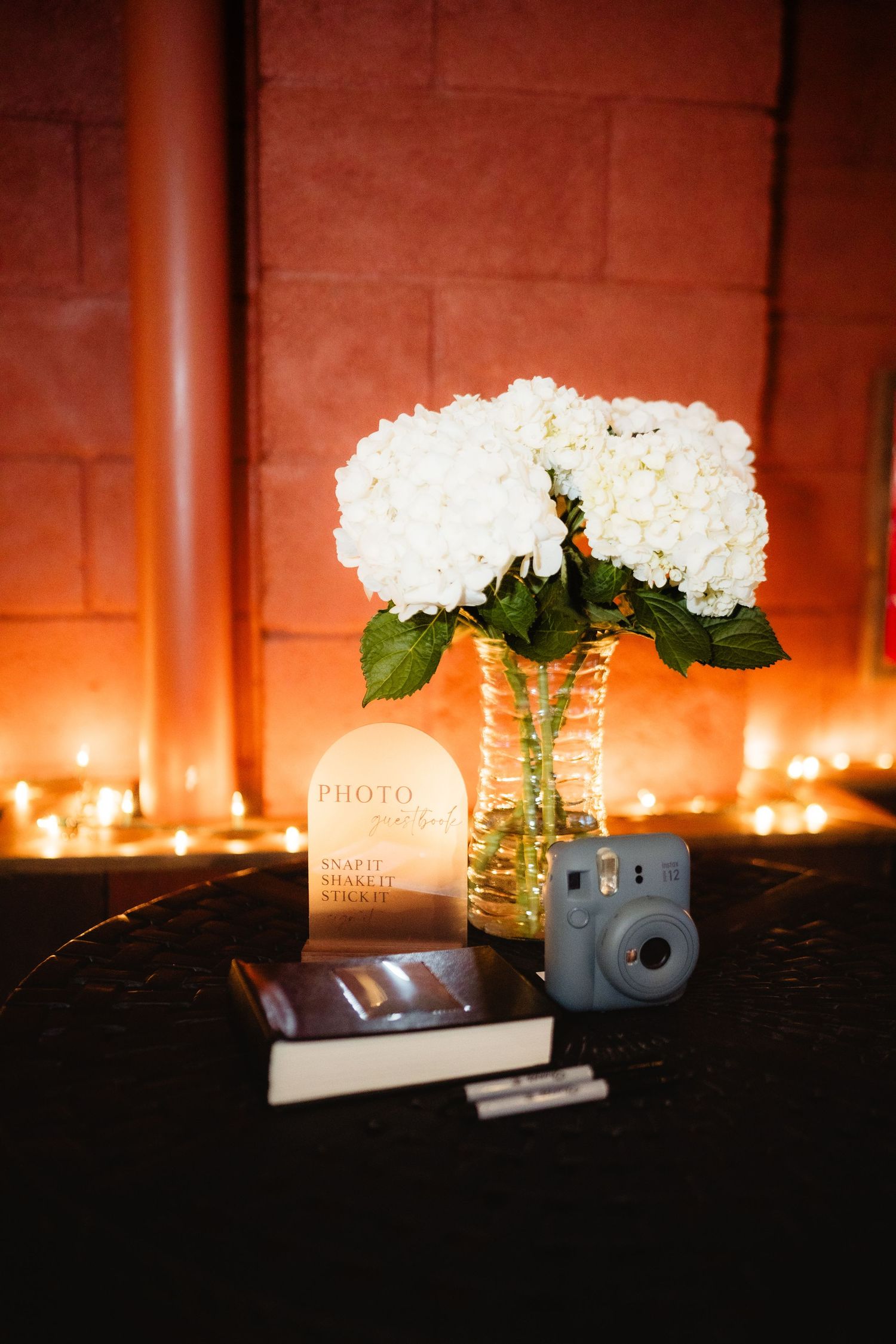 White hydrangeas in gold vase with Instax camera, sign, and fairy lights on dark table at wedding reception.