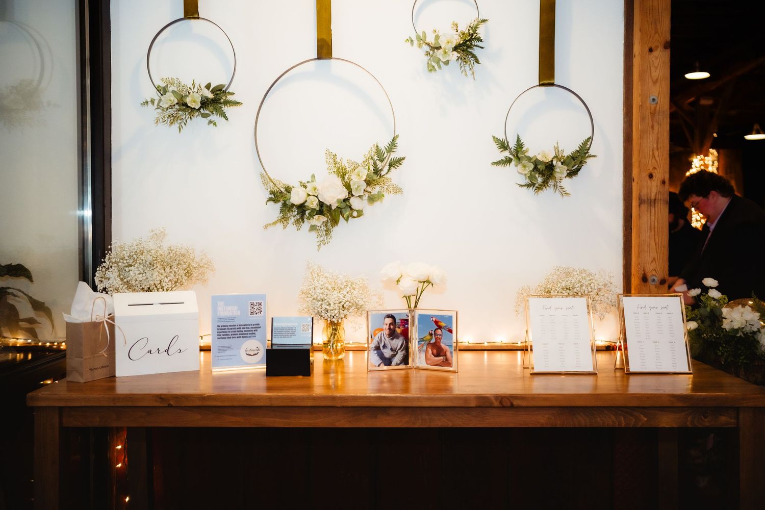 Elegant wedding reception table with floral hoop wreaths, baby's breath, framed photos, and signage on rustic wood.