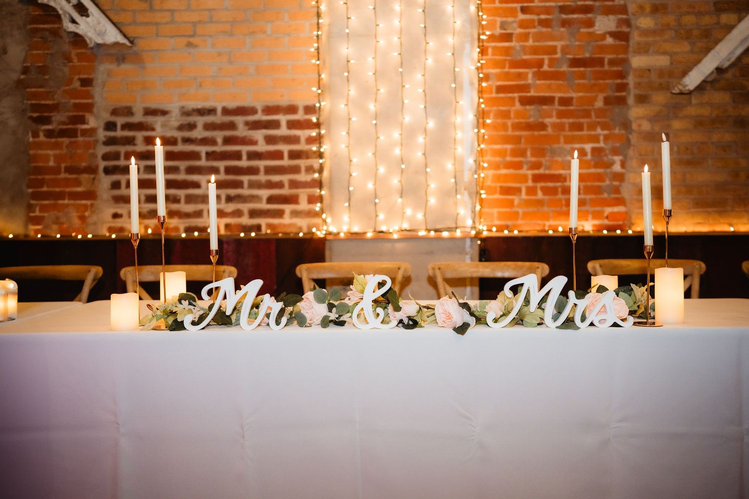 Elegant wedding head table with Mr. & Mrs. signs, white candles, and fairy lights against exposed brick wall.