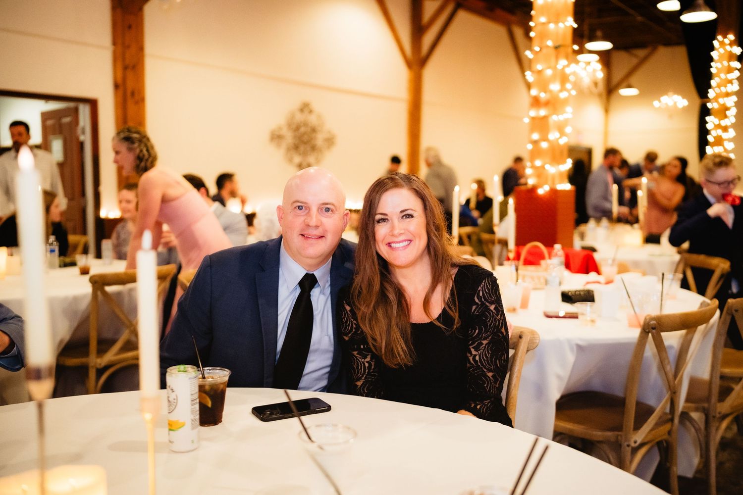 Couple smiling together at a wedding reception dinner table in an elegant venue with warm string lights.