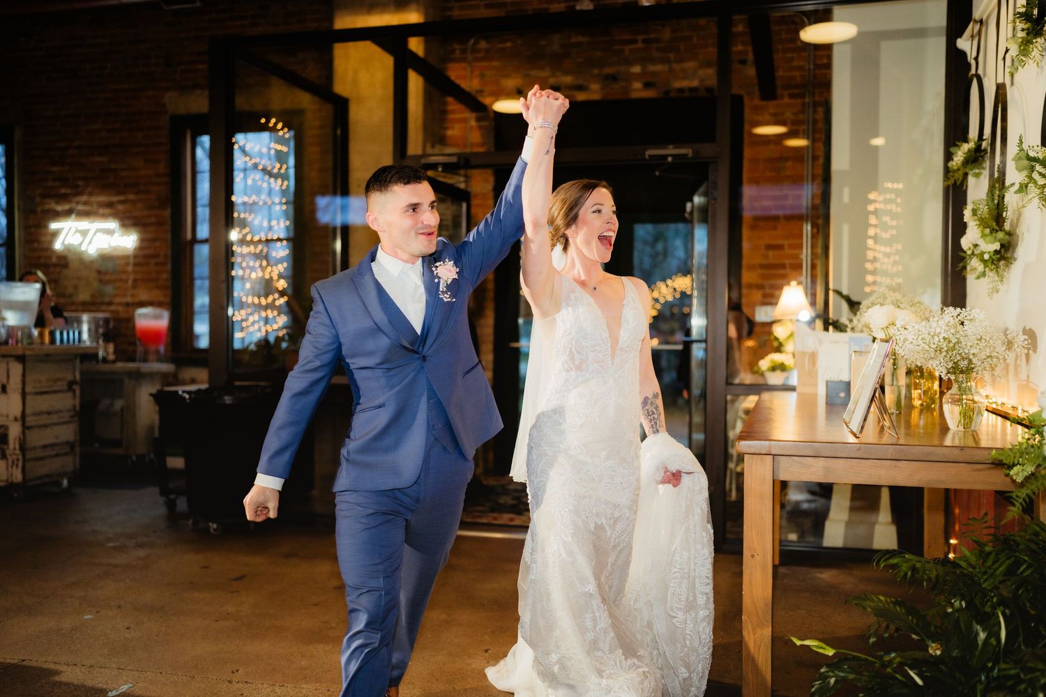 Bride and groom celebrate their grand entrance at a rustic wedding venue, raising hands joyfully in a blue suit and lace gown.