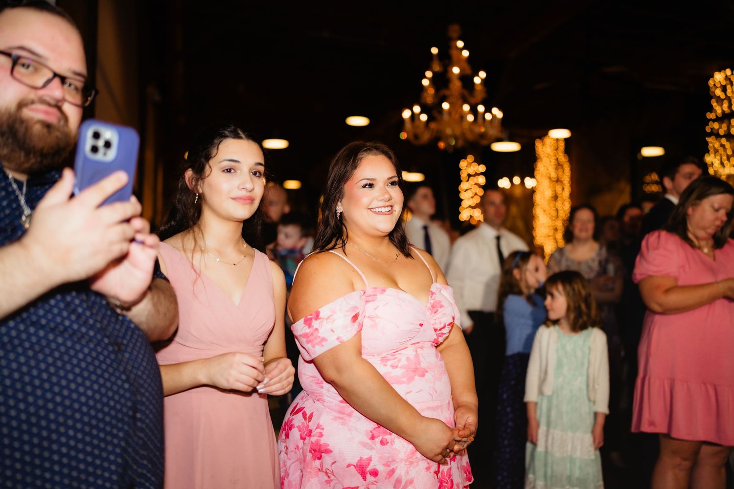 Two women in pink dresses smile at a wedding reception with warm chandelier lighting in the background.