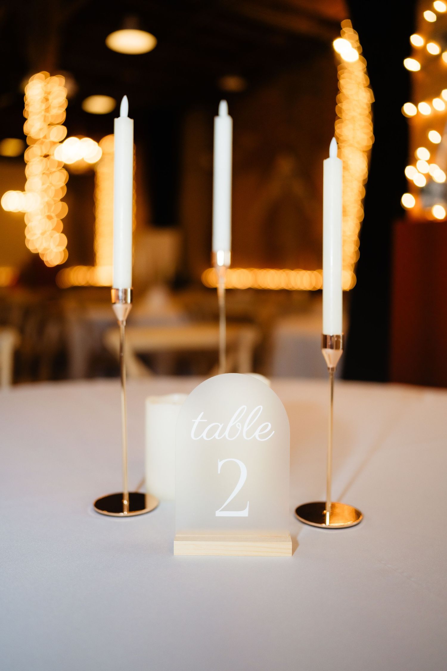 Elegant wedding table setting with gold candlesticks, white taper candles, and illuminated acrylic table number 2 sign.