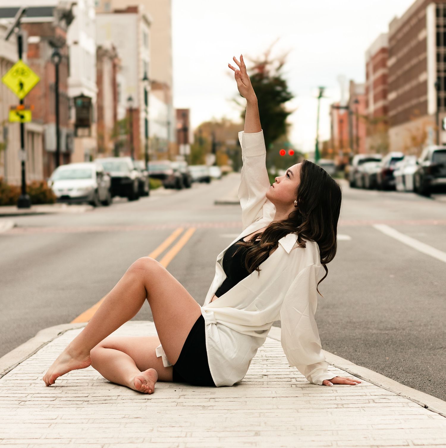 A barefoot woman in black shorts and white shirt poses gracefully on a city street median, arm raised elegantly.