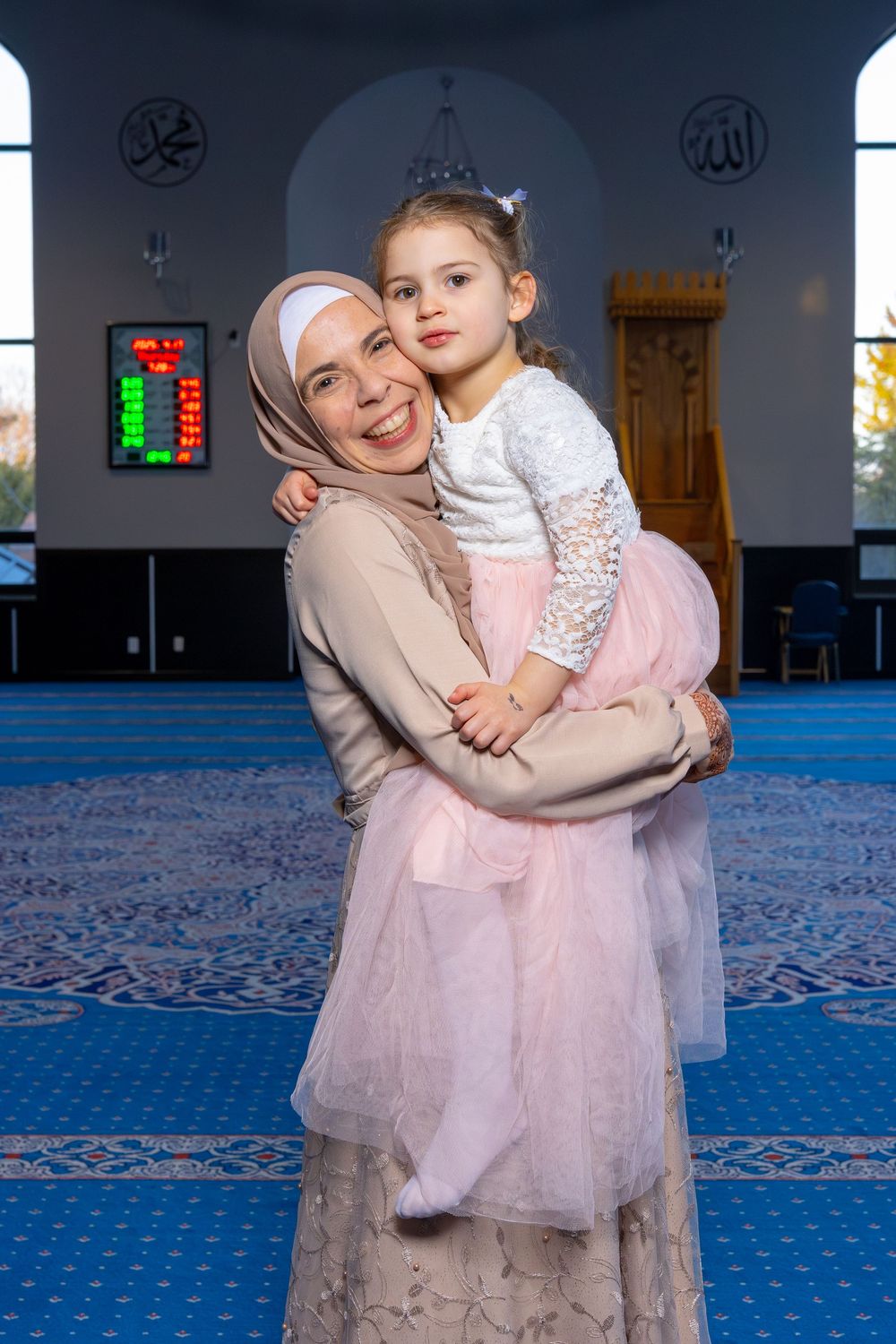 Muslim mother in hijab holding young daughter in pink tutu dress inside a mosque with blue carpet.