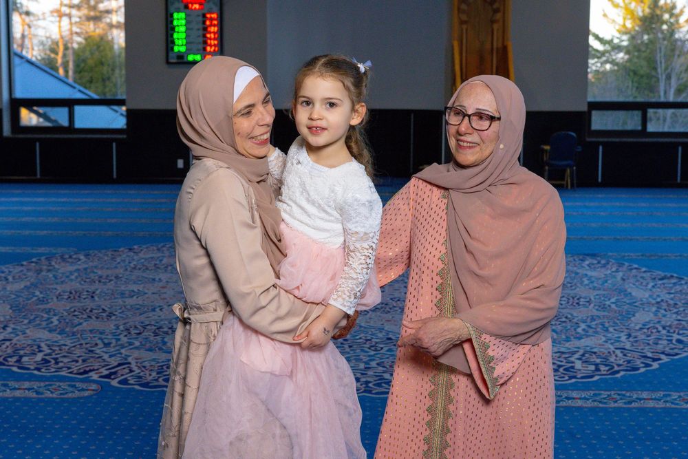 Two women wearing hijabs smile while holding a young girl in a decorated mosque interior.