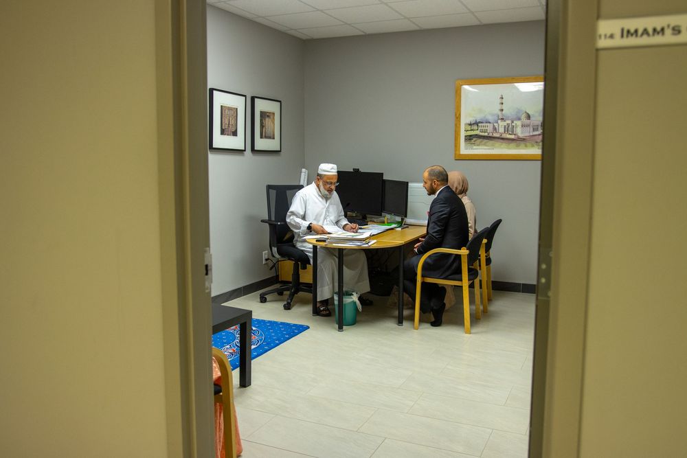 Imam meeting with a visitor in his office inside a mosque, viewed through a doorway.