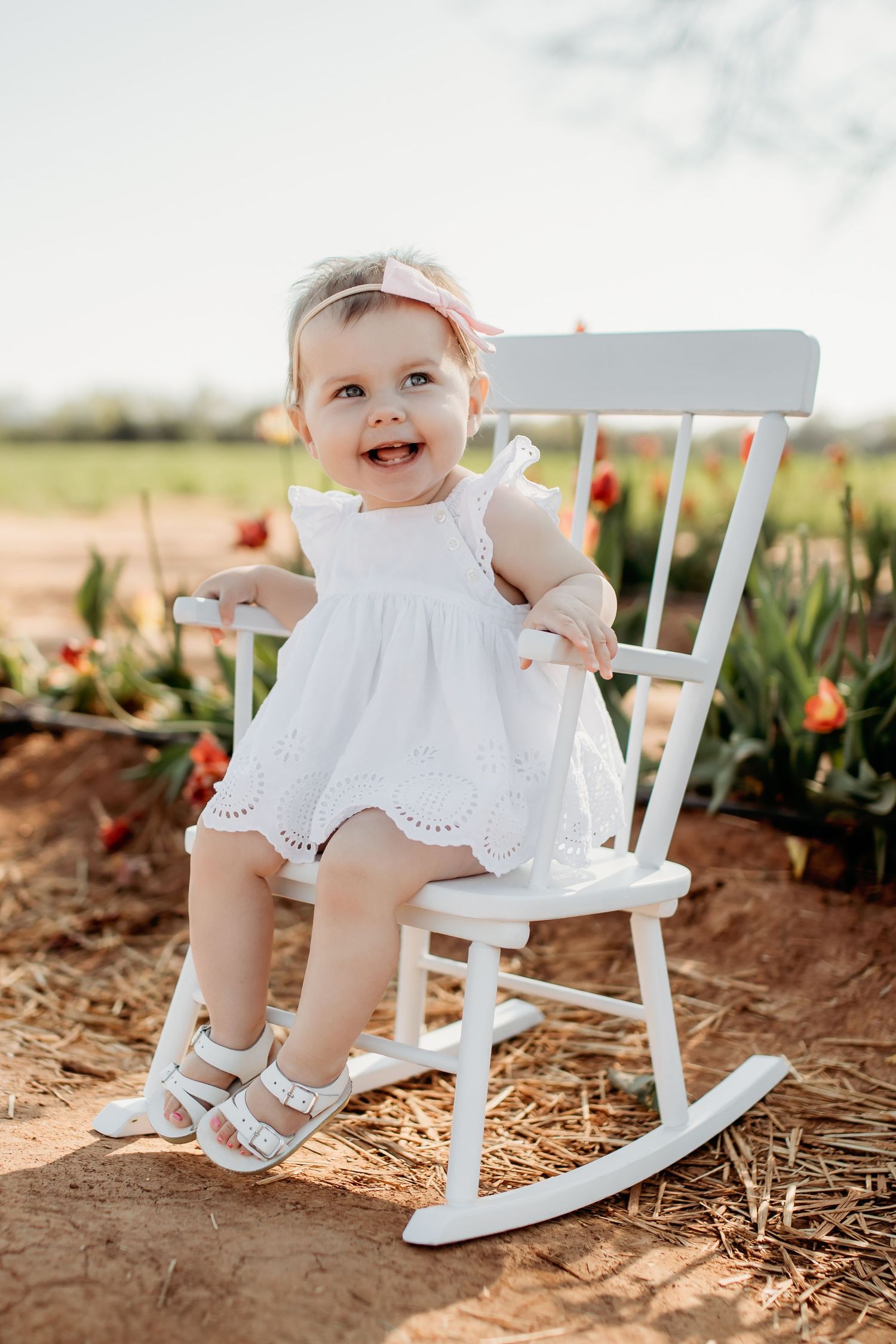 Three photos show a baby in a white dress sitting on a small white rocking chair in a garden with tulips.