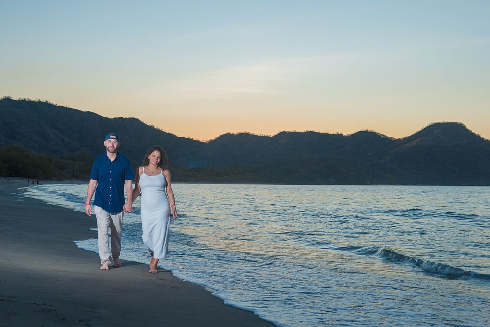 Couple walking together on a scenic beach at sunset with mountains and ocean waves in the background.